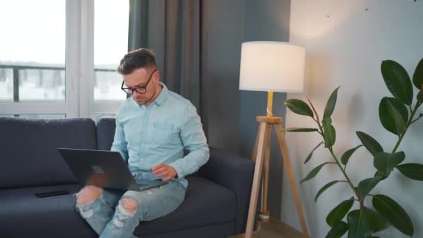 戴眼镜的男人坐在沙发上，带着笔记本电脑，在舒适的房间里工作。办公室外的远程工作. — 图库视频影像