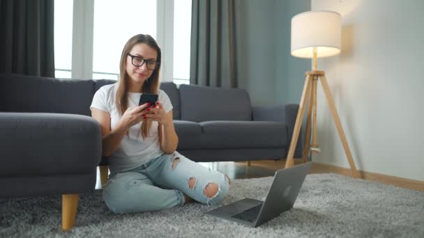 Lässig gekleidete Frau sitzt auf Teppich mit Laptop und Smartphone und arbeitet in gemütlichem Raum. Fernarbeit außerhalb des Büros. — Stockvideo