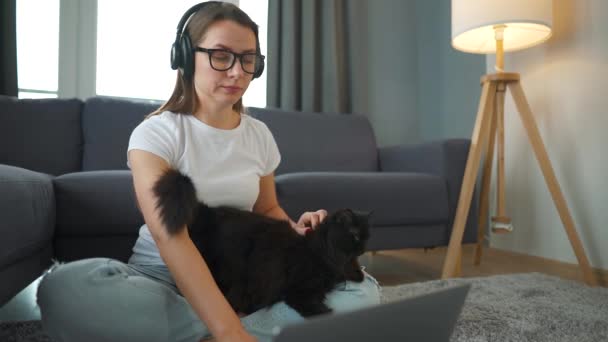 Casually klädd kvinna sitter på en matta med en bärbar dator, håller på knäna och stryker en fluffig katt och arbetar i ett mysigt rum. Fjärrarbete utanför kontoret. — Stockvideo