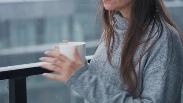 Donna caucasica rimane sul balcone durante la nevicata con una tazza di caffè caldo o tè. Guarda i fiocchi di neve e respira l'aria fresca gelida — Video Stock