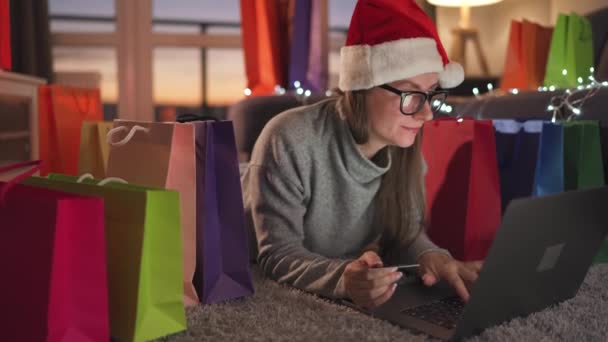 La donna con gli occhiali che indossa un cappello di Babbo Natale è sdraiata sul tappeto ed effettua un acquisto online utilizzando una carta di credito e un laptop. Shopping bag in giro. — Video Stock