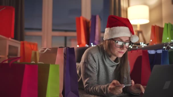 Mujer con gafas con un sombrero de Santa Claus está acostado en la alfombra y hace una compra en línea con una tarjeta de crédito y un ordenador portátil. Bolsas de compras alrededor. — Vídeos de Stock