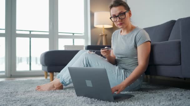 Woman with glasses sits on the floor and makes an online purchase using a credit card and laptop. Online shopping, lifestyle technology — Stock Video