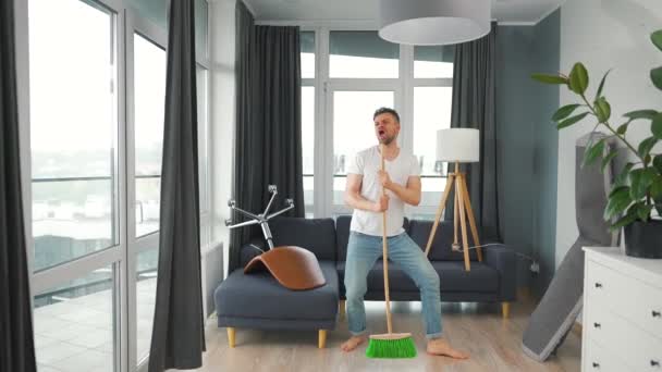 Homem limpando a casa e se divertindo dançando e cantando com uma vassoura. Movimento lento — Vídeo de Stock