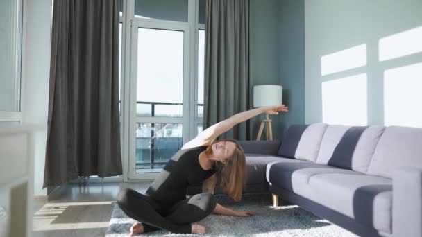 Jonge blanke vrouw in zwarte jumpsuit doet yoga thuis om flexibiliteit en evenwicht te ontwikkelen. Langzame beweging — Stockvideo