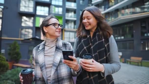 Două femei fericite mergând cu cafeaua takeaway și vorbind cu interes între ele în cartierul de afaceri . — Videoclip de stoc