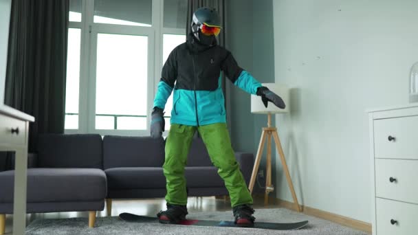 Un vídeo divertido. Hombre vestido de snowboarder representa el snowboard en una alfombra en una habitación acogedora. Esperando un invierno nevado. Movimiento lento — Vídeos de Stock