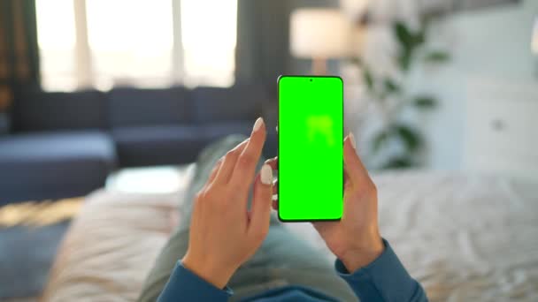 Kvinna hemma liggande på en säng och med hjälp av smartphone med grön mock-up skärm i vertikalt läge. Flicka surfa på Internet, titta på innehåll, videor. Födelsedatum. — Stockvideo