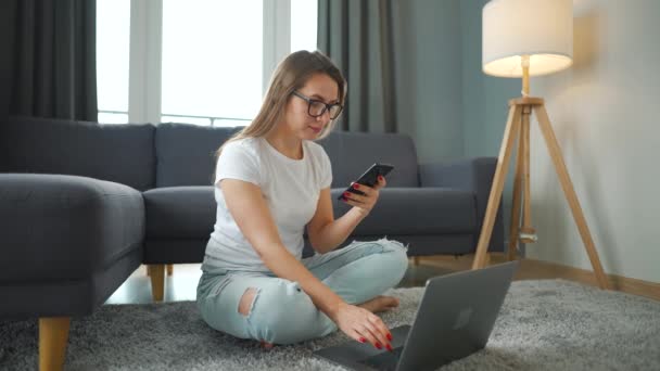 Halıda dizüstü bilgisayarı ve akıllı telefonu olan rahat bir odada çalışan sıradan giyinmiş bir kadın. Ofis dışında uzaktan çalışma. — Stok video