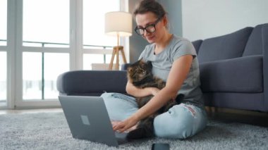 Sıradan giyimli bir kadın dizüstü bilgisayarla halıya oturur, dizlerinin üzerine çöker ve tüylü bir kediyi okşar ve rahat bir odada çalışır. Ofis dışında uzaktan çalışma.