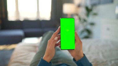 Evdeki kadın yatakta uzanıyor ve dikey modda yeşil maket ekranlı akıllı telefon kullanıyor. Kız internette geziniyor, içeriği izliyor, videolar izliyor. POV.