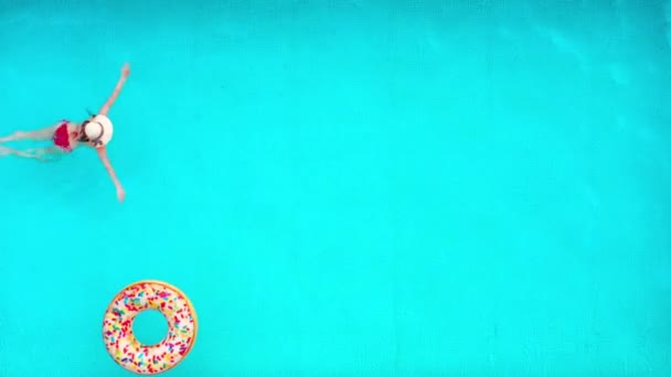 Blick von oben, als eine Frau im roten Badeanzug im Pool schwimmt. Bunte aufblasbare Donuts schweben in ihrer Nähe. Sommerlicher Lebensstil — Stockvideo