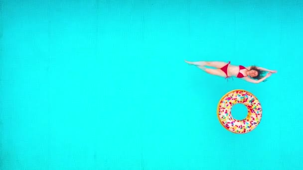 Blick von oben auf eine Frau im roten Badeanzug, die auf dem Rücken im Pool liegt. Bunte aufblasbare Donuts schweben in ihrer Nähe. Entspanntes Konzept — Stockvideo