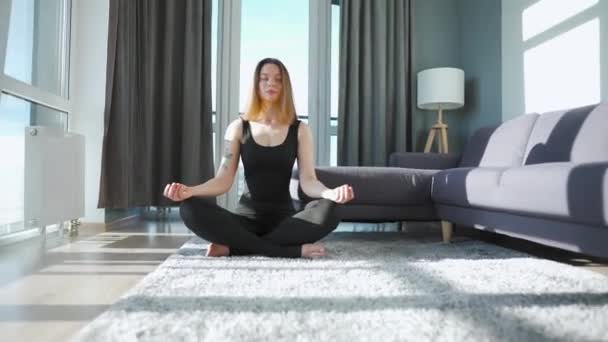 Mujer joven caucásica en mono negro sentada en posición de loto y meditando, yoga en casa. Movimiento lento — Vídeo de stock
