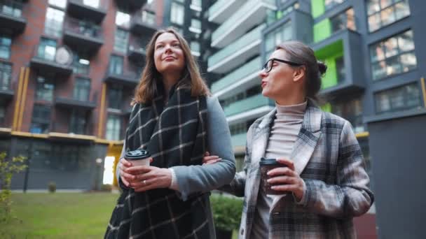 Zwei fröhliche Frauen spazieren mit Kaffee zum Mitnehmen durch das Geschäftsviertel und unterhalten sich interessiert untereinander. Zeitlupe — Stockvideo