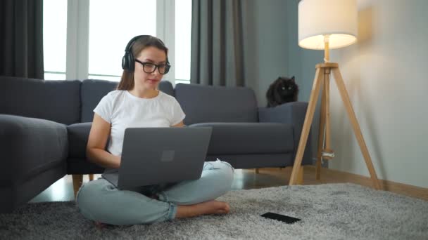 Casually klädd kvinna med hörlurar sitter på matta med laptop och arbetar i mysiga rum. Fjärrarbete utanför kontoret. — Stockvideo