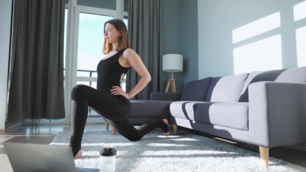 Jonge blanke vrouw in zwarte jumpsuit doet yoga thuis om flexibiliteit en evenwicht te ontwikkelen op de video-tutorial op het internet. Langzame beweging — Stockvideo