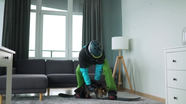 Un vídeo divertido. Hombre vestido de snowboarder monta una tabla de snowboard en una alfombra en una habitación acogedora. Tiene un gato esponjoso en sus brazos. Esperando un invierno nevado. Movimiento lento — Vídeos de Stock