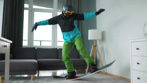 Un vídeo divertido. Hombre vestido de snowboarder representa el snowboard en una alfombra en una habitación acogedora. Esperando un invierno nevado. Movimiento lento — Vídeos de Stock