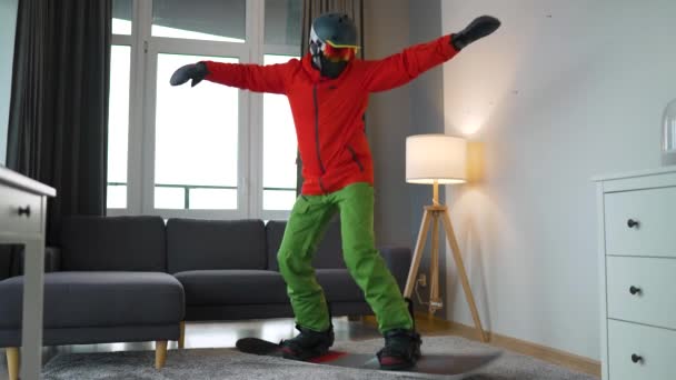 Divertente video. L'uomo vestito da snowboarder raffigura lo snowboard su un tappeto in una stanza accogliente. Aspettando un inverno nevoso. — Video Stock