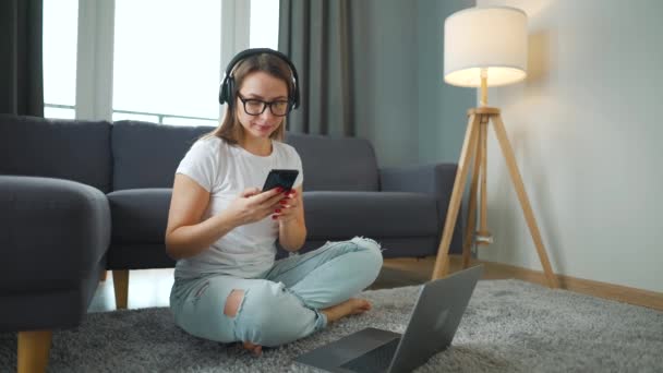 Kulaklıklı sıradan giyinmiş bir kadın halının üzerinde oturuyor, akıllı telefon ve dizüstü bilgisayarla ve rahat bir odada çalışıyor. Ofis dışında uzaktan çalışma. — Stok video