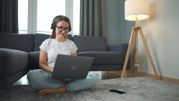 Casualmente mujer vestida con auriculares está sentado en la alfombra con el ordenador portátil y trabajando en una habitación acogedora. Trabajo remoto fuera de la oficina. — Vídeos de Stock