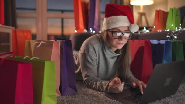 Frau mit Brille und Weihnachtsmannmütze liegt auf dem Teppich und kauft online mit Kreditkarte und Laptop ein. Einkaufstüten herum. — Stockvideo