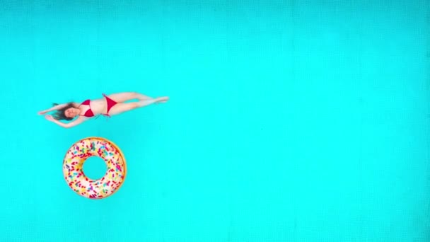 Blick von oben auf eine Frau im roten Badeanzug, die auf dem Rücken im Pool liegt. Bunte aufblasbare Donuts schweben in ihrer Nähe. Entspanntes Konzept — Stockvideo