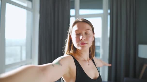 Молода біла жінка в чорному комбінезоні робить йогу вірабхадрасану вдома, щоб розвинути гнучкість і рівновагу. Повільний рух — стокове відео