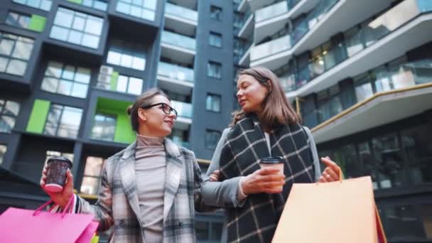 Två glada kvinnor gå med shoppingväskor och hämtmat kaffe efter en lyckad shopping och prata med intresse sinsemellan. Långsamma rörelser — Stockvideo