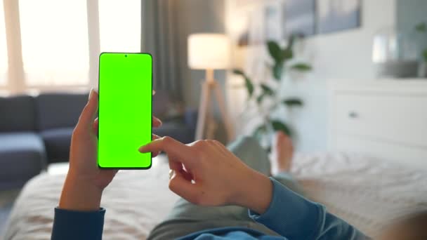 Evdeki kadın yatakta uzanıyor ve dikey modda yeşil maket ekranlı akıllı telefon kullanıyor. Kız internette geziniyor, içeriği izliyor, videolar izliyor. POV. — Stok video