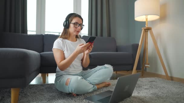 Mulher casualmente vestida com fones de ouvido está sentado no tapete com smartphone e laptop e trabalhando em quarto acolhedor. Trabalho remoto fora do escritório. — Vídeo de Stock