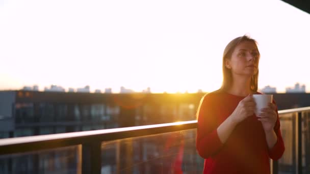 Mulher com uma xícara de café em pé na varanda e admirar o pôr do sol — Vídeo de Stock