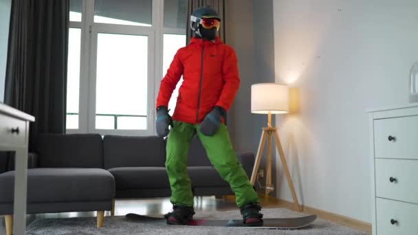 Vtipné video. Muž oblečený jako snowboardista šťastně tančí a baví se, zatímco stojí na snowboardu na koberci v útulné místnosti. Čekání na sněhovou zimu. — Stock video