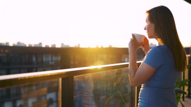 Kobieta z filiżanką kawy stojąca na balkonie i podziwiająca zachód słońca — Wideo stockowe