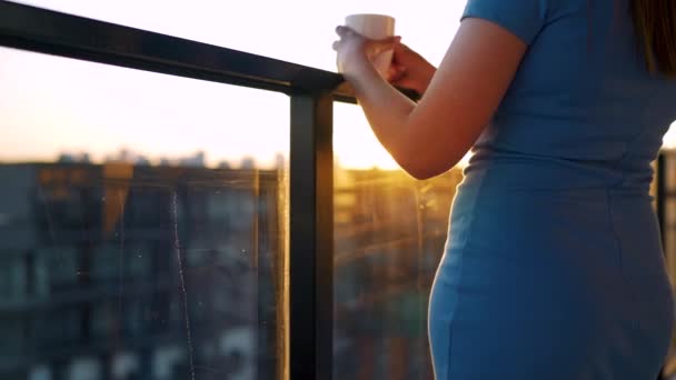 Mulher com uma xícara de café em pé na varanda e admirar o pôr do sol — Vídeo de Stock