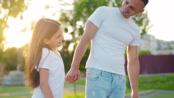 Apa és lánya sétálnak a környéken napnyugtakor. A gyerek fogja az apák kezét. Lassú mozgás. — Stock videók