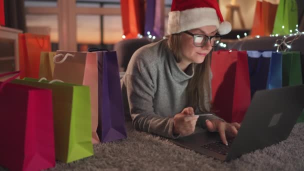 La donna con gli occhiali che indossa un cappello di Babbo Natale è sdraiata sul tappeto ed effettua un acquisto online utilizzando una carta di credito e un laptop. Shopping bag in giro. — Video Stock