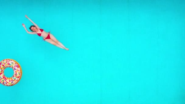 プールに横たわっている赤い水着の女性として上からの眺め。多色のインフレータブルドーナツが彼女の近くに浮かぶ。リラックスしたコンセプト — ストック動画