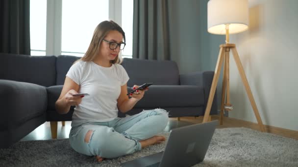 Mujer con gafas se sienta en el suelo y hace una compra en línea con una tarjeta de crédito y un teléfono inteligente. Compras en línea, tecnología de estilo de vida — Vídeos de Stock