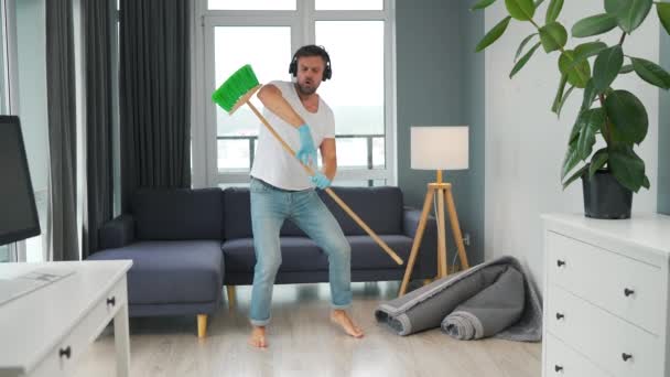 El hombre con auriculares hace la limpieza y se imagina a sí mismo como una estrella de rock, toca la escoba como una guitarra. Movimiento lento — Vídeos de Stock