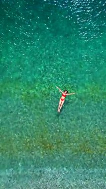 Kırmızı mayo giymiş bir kadının hava görüntüsü, kristal berrak deniz sularında rahatlama somutlaştırıcı bir şekilde sırtüstü uzanır. Yaz tatilleri. Dikey video