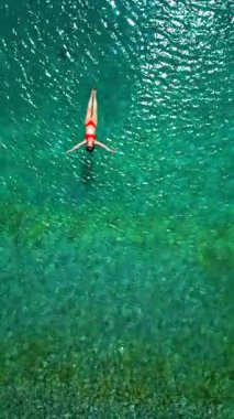 Kırmızı mayo giymiş bir kadının hava görüntüsü, kristal berrak deniz sularında rahatlama somutlaştırıcı bir şekilde sırtüstü uzanır. Yaz tatilleri. Dikey video