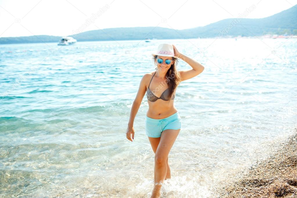 Young woman in a hat and swimsuit walking on the beach on a hot