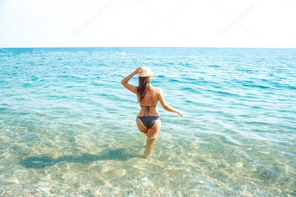 Woman in sun hat and bikini standing with her arm raised to her