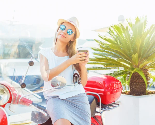 Happy young woman drinking takeaway coffee near her red moped in — Zdjęcie stockowe