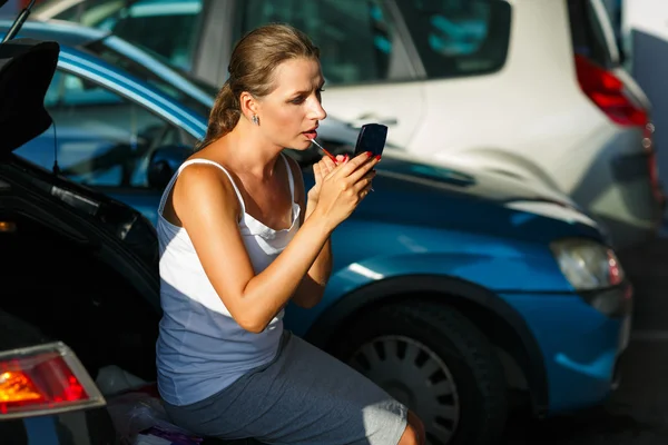 Mujer joven pintarse los labios sentado en el maletero de un coche en el — Foto de Stock