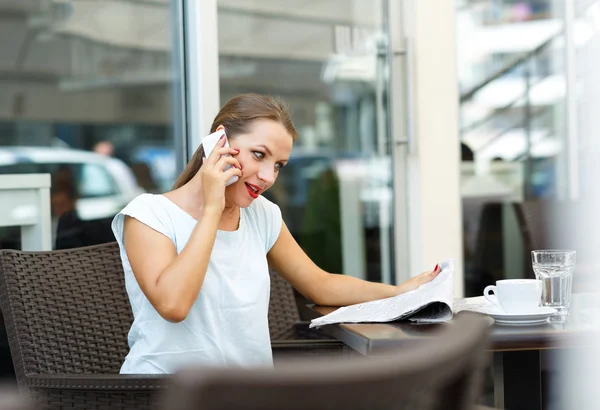 Young woman reading a newspaper and talking on a cell phone sitt — Stock Photo, Image