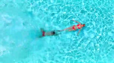 Çift havuzda rahatlar. Kadın sırtüstü yatarken, bir erkeğin havuza dalıp yüzdüğü tepeden aşağı manzara..