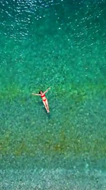 Kırmızı mayo giymiş bir kadının hava görüntüsü, kristal berrak deniz sularında rahatlama somutlaştırıcı bir şekilde sırtüstü uzanır. Yaz tatilleri. Dikey